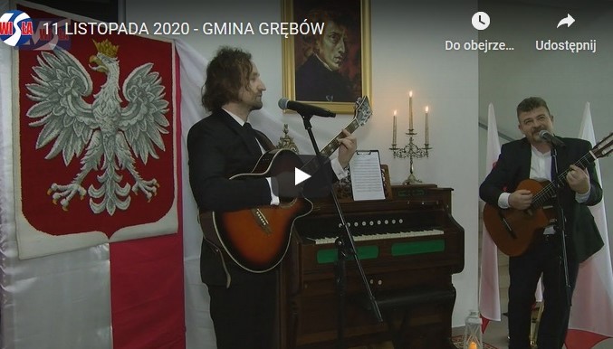 Dwóch mężczyzn z gitarami w ręku stoją przed mikrofonami. Za mini znajduje się fortepian oraz godło Polski (biały orzeł na czerwonym tle)