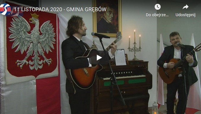 Dwóch mężczyzn z gitarami w ręku stoją przed mikrofonami. Za mini znajduje się fortepian oraz godło Polski (biały orzeł na czerwonym tle)