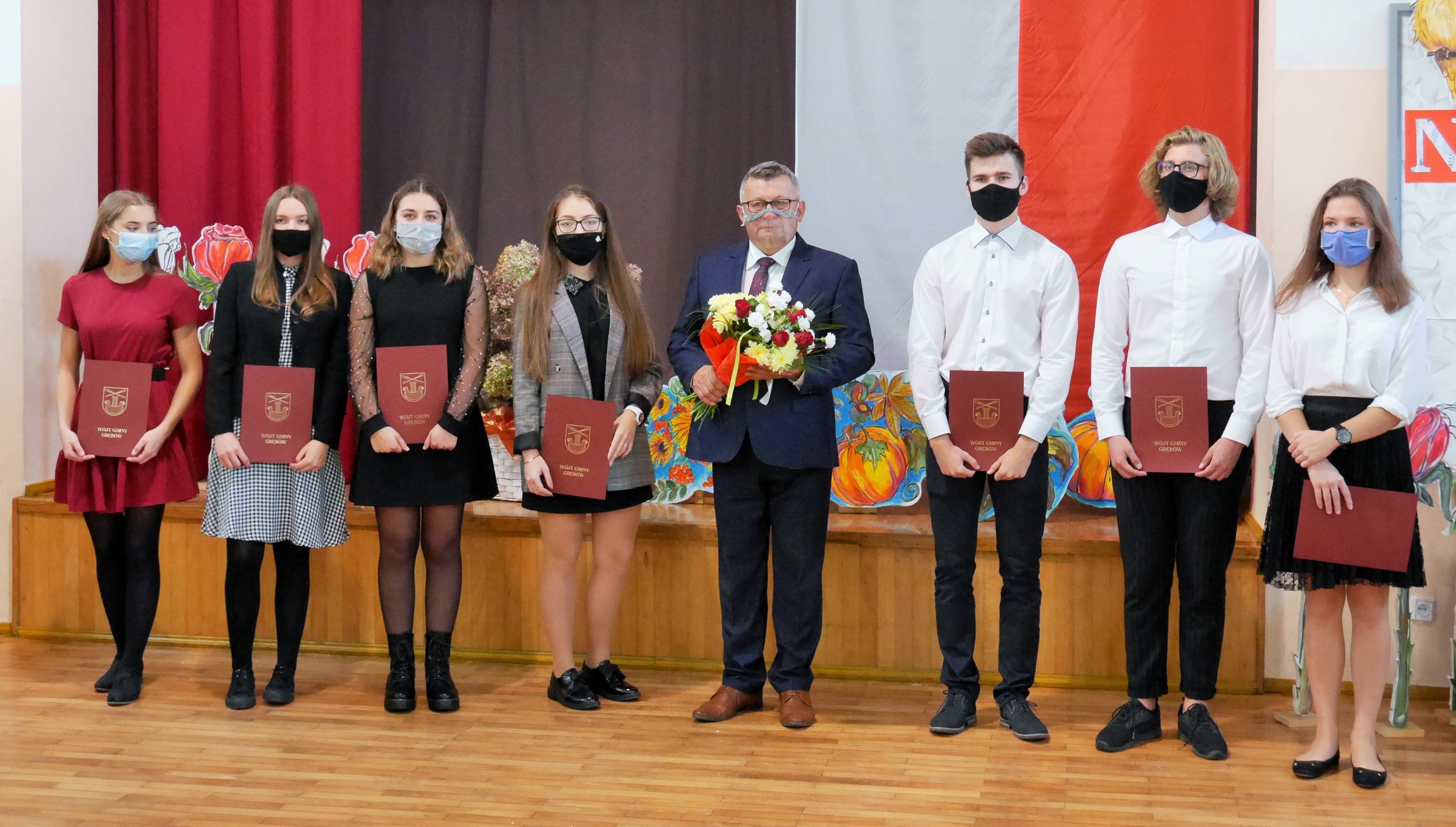 W pomieszczeniu stoi w rzędzie grupa młodzieży w maseczkach na twarzy. W ręku trzymają bordowe teczki. Pośród nich mężczyzna w garniturze z bukietem kwiatów w rękach. 