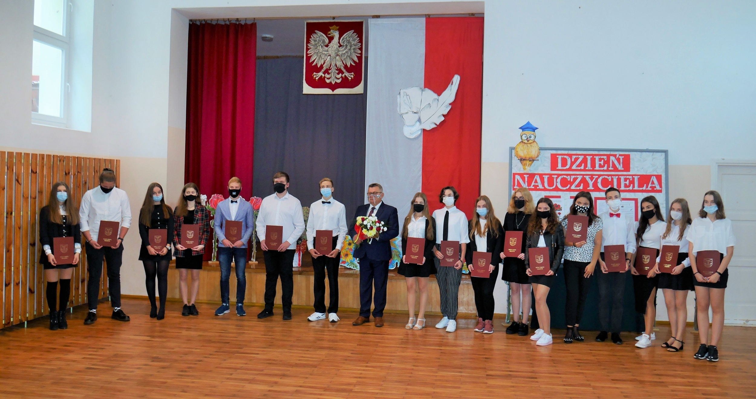 W pomieszczeni stoi w rzędzie liczna grupa młodzieży w maseczkach na twarzy. W ręku trzymają bordowe teczki. Pośród nich mężczyzna w garniturze z bukietem kwiatów w rękach. 