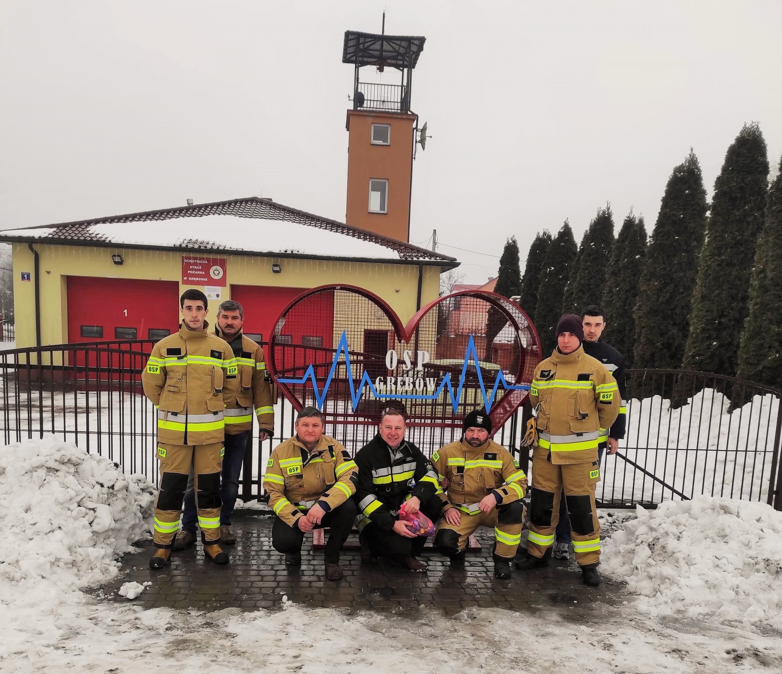 Grupa strażaków stoi obok metalowego serca na nakrętki. W tle remiza strażacka. 