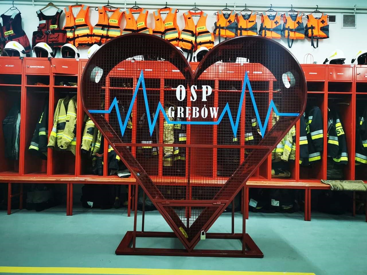 Duże, czerwone, metalowe serce (pojemnik) z napisem OSP Grębów. W tle szafki z ubraniami, butami strażaków