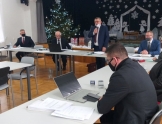 Grupa osób siedzących przy stołach. Jeden mężczyzna stoi i trzyma mikrofon w ręce. W tle widoczna choinka i dekoracja świąteczna.