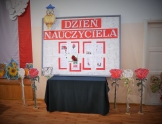 Pozioma tablica stojąca na stole przykrytym ciemnym materiałem z napisem dzień nauczyciela . W lewym roku ozdobna sowa w  niebieskiej czapce. Na podłodze stoją dekoracyjne białe i czerwone kwiaty.