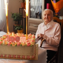 Starsza kobieta siedzi na krześle. Przed nią na stole leży tort ze świeczkami 100 
