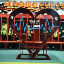 Duże, czerwone, metalowe serce (pojemnik) z napisem OSP Grębów. W tle szafki z ubraniami, butami strażaków