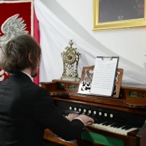 Mężczyzna gra na fortepianie, na który stoi kartka z nutami oraz świecznik. Po prawej stronie widoczny godło Polski (biały orzeł na czerwonym tle)